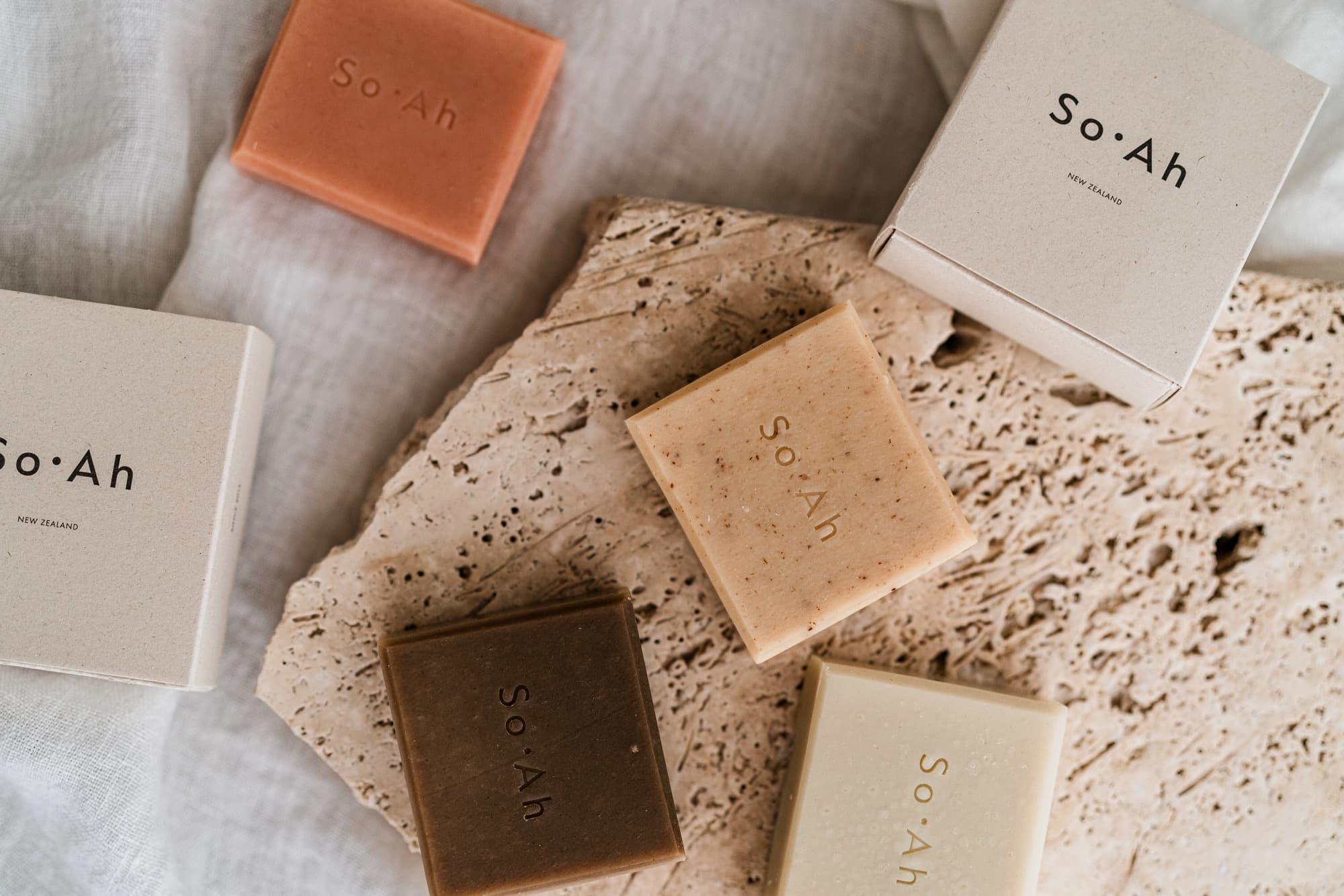 So·Ah New Zealand soap bars in earthy tones arranged with their kraft packaging on a travertine slab and linen.