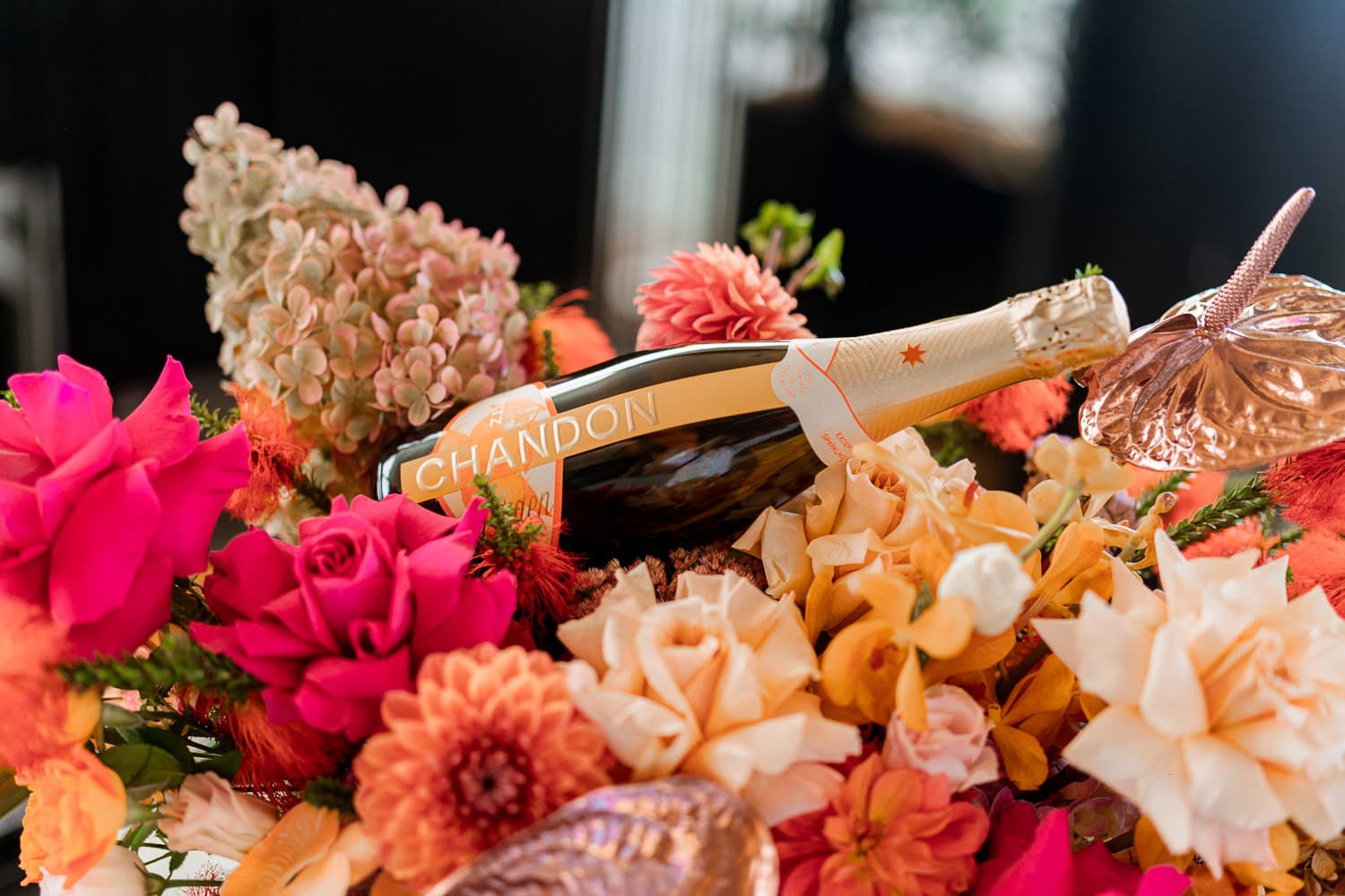 A bottle of Chandon sparkling wine nestled in a lush arrangement of pink and coral roses, dahlias and hydrangeas.