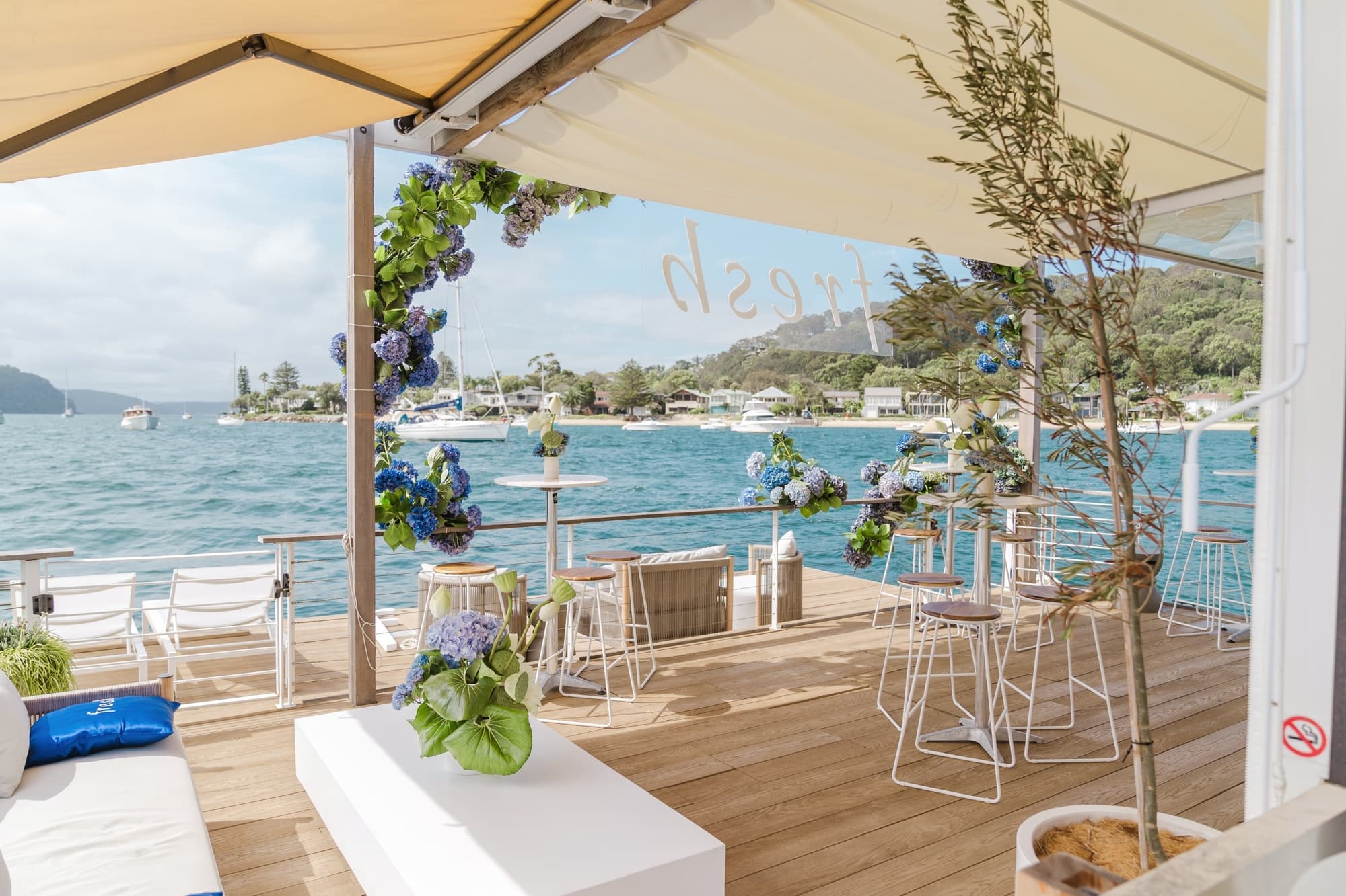 A waterfront event deck with white bar stools and blue hydrangeas overlooking a boat-dotted bay, 'Fresh' lettering on the glass.