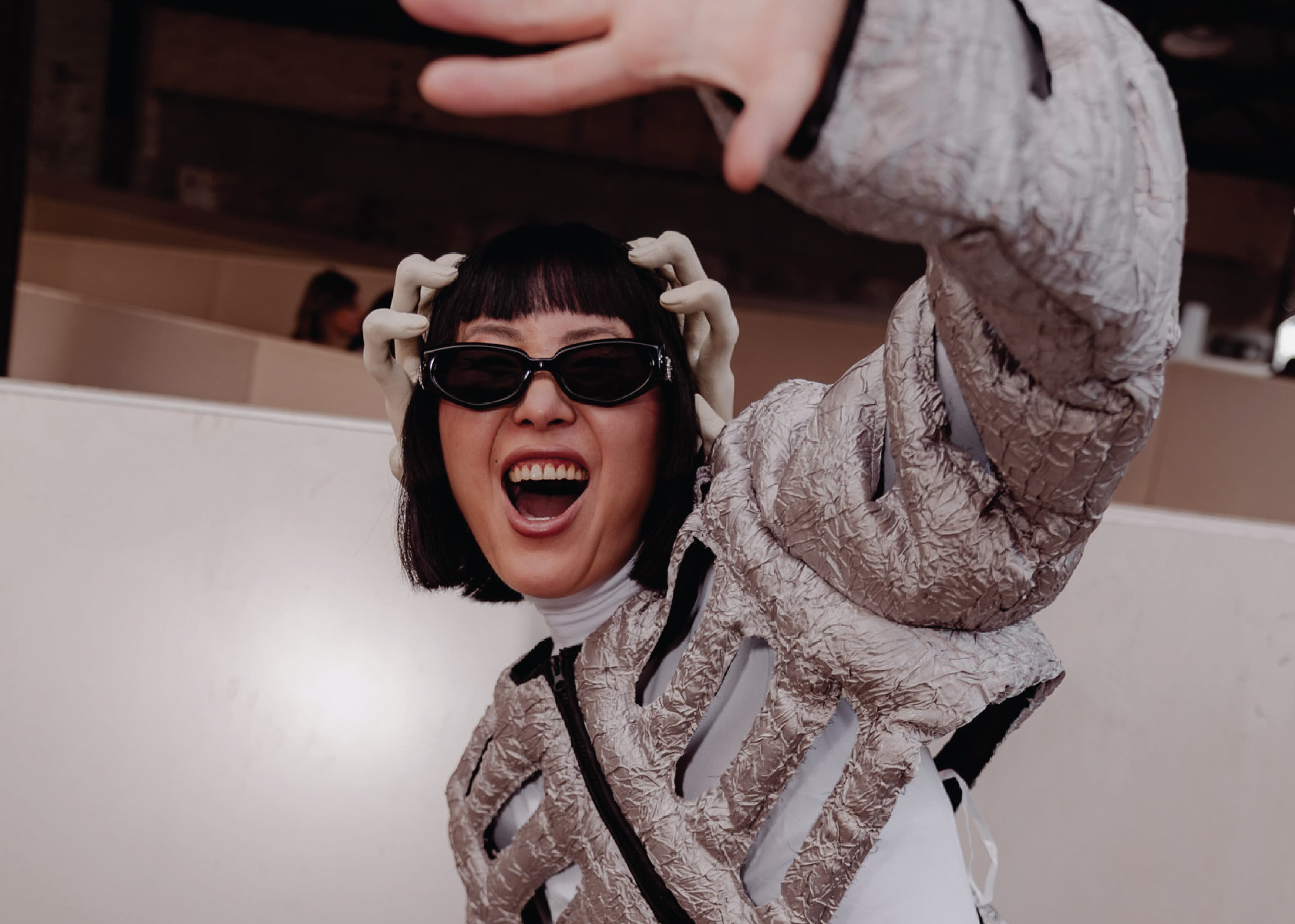 Street-style guest in a crinkled silver metallic jacket and black sunglasses laughs and throws a hand toward the camera at a fashion event.