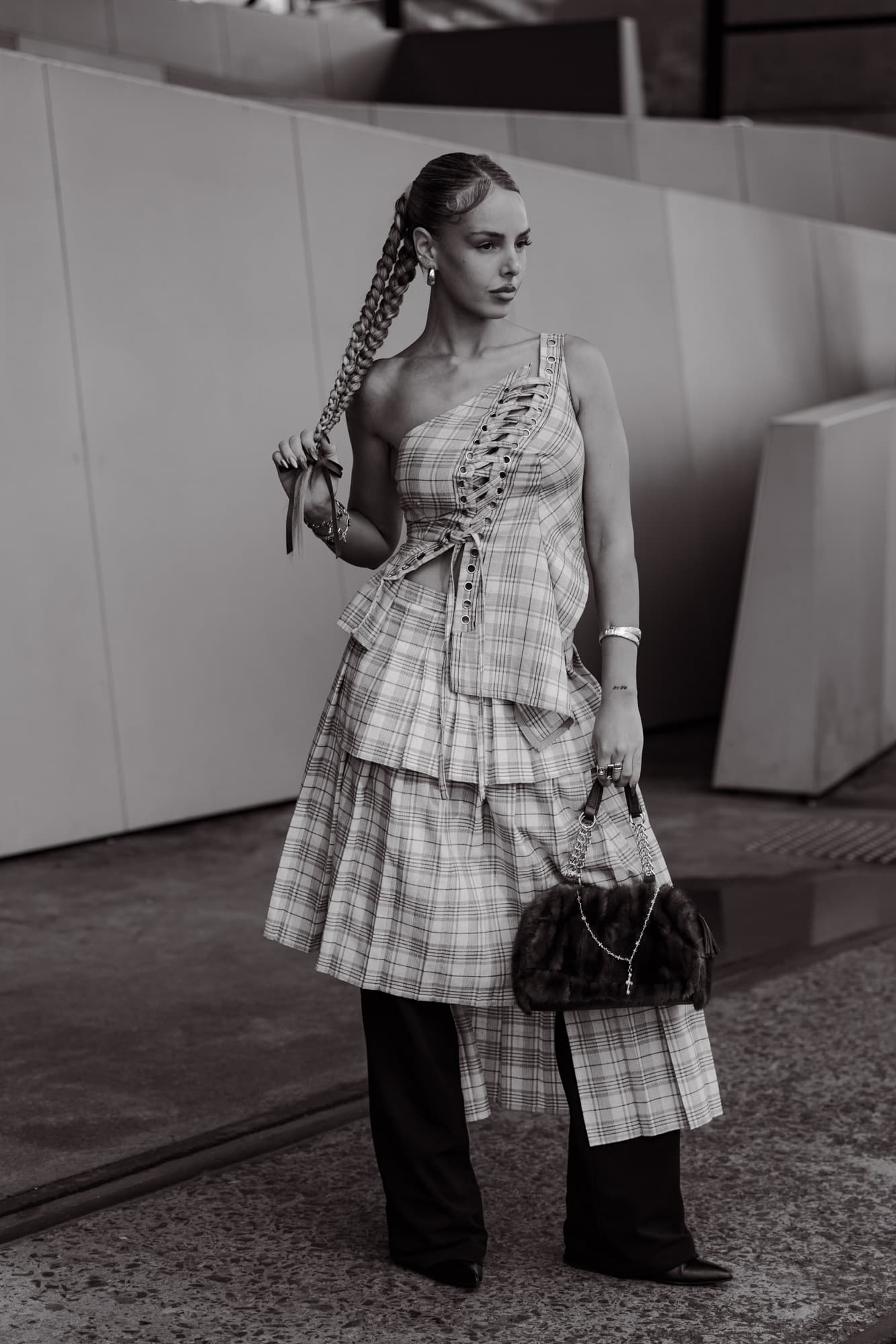 Black-and-white street-style portrait of a guest in a plaid one-shoulder tiered dress with a long braid, holding a fur-trimmed bag.