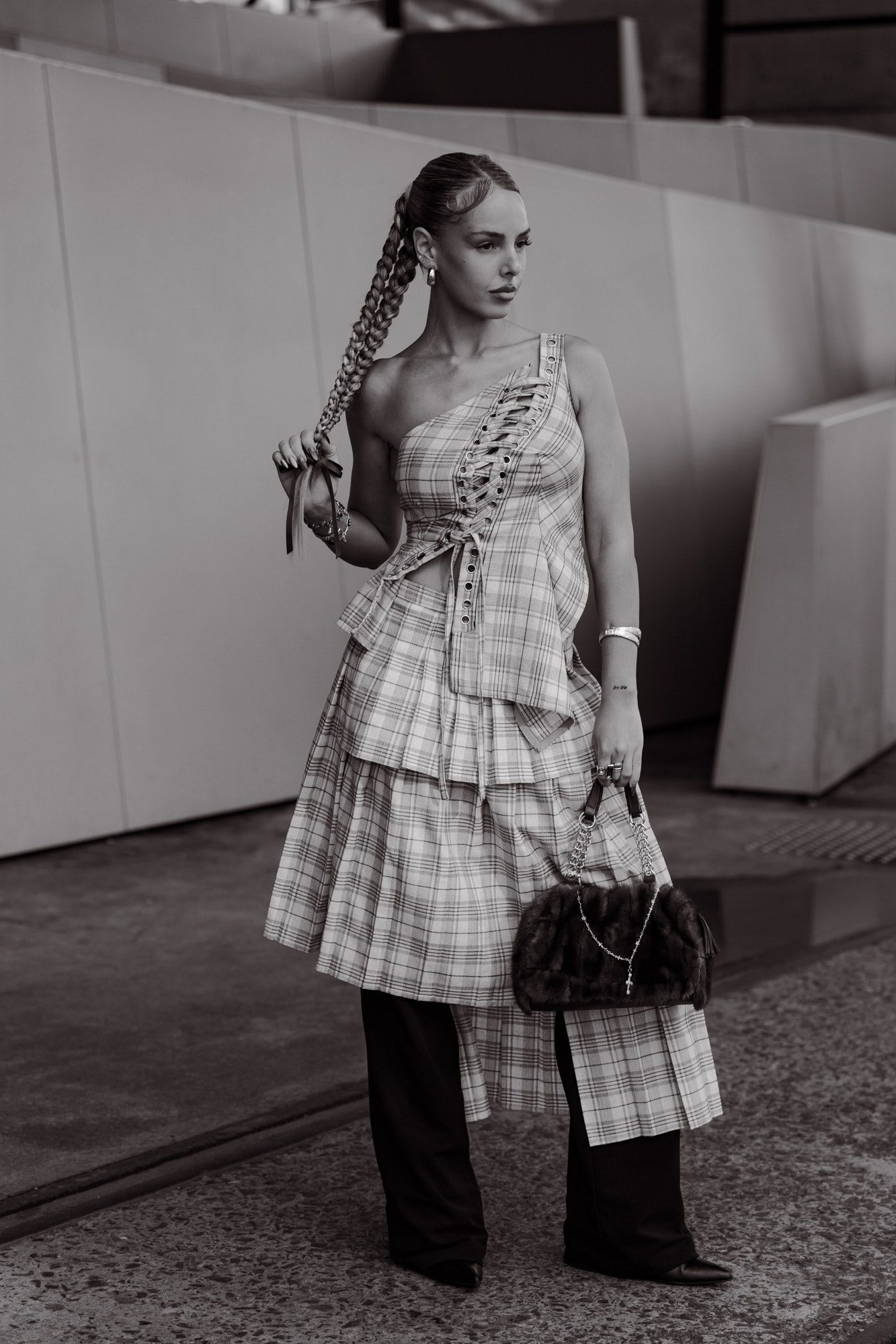 Black-and-white street-style portrait of a guest in a plaid one-shoulder tiered dress with a long braid, holding a fur-trimmed bag.