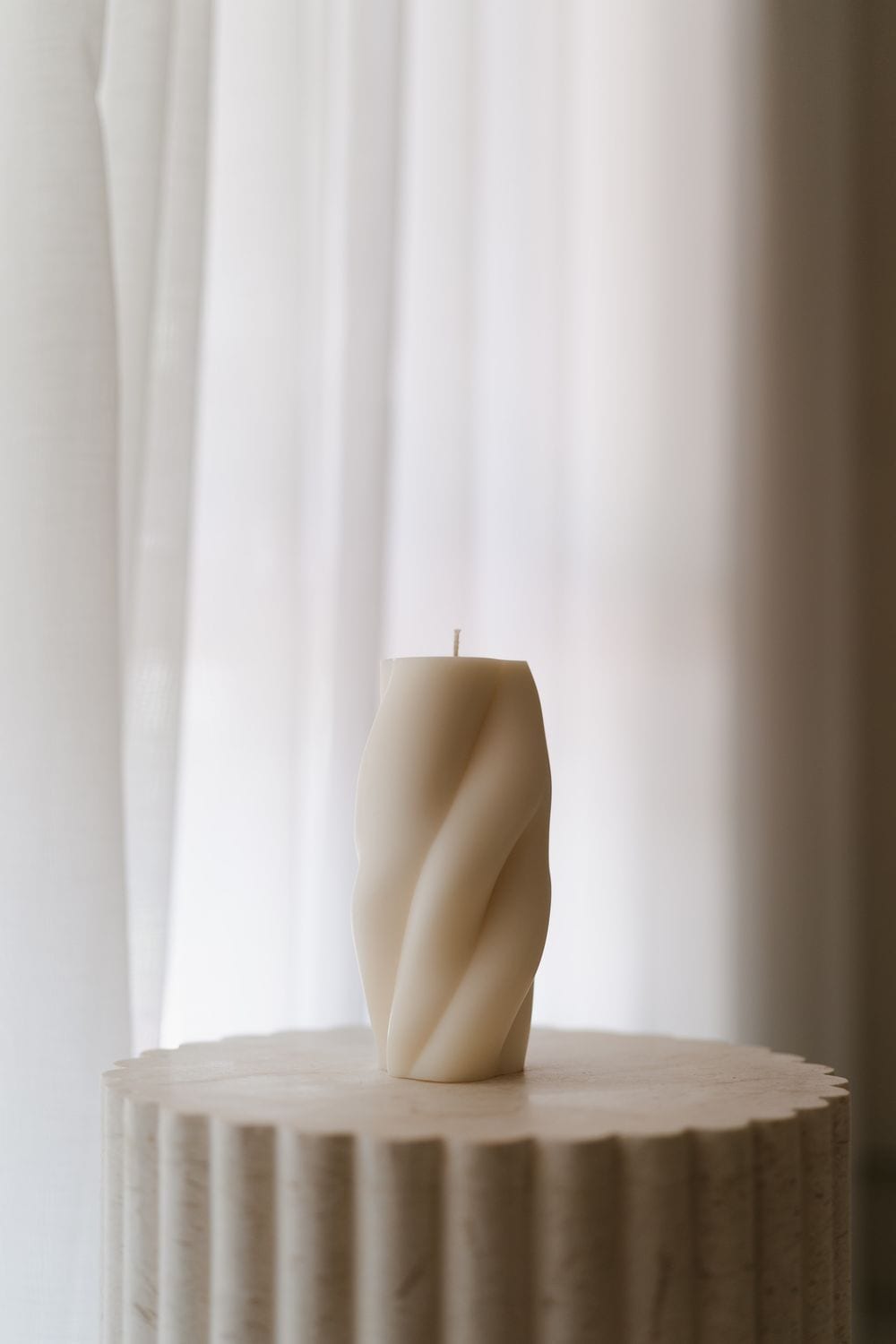 Cream twisted sculptural pillar candle standing on a fluted travertine pedestal against soft sheer curtains.